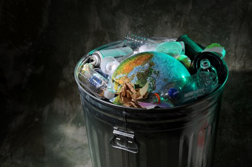 A metallic grey, cylindrical rubbish bin with a closed lid, positioned outdoors on a grey paving surface. The bin is overfilled with various types of waste, including numerous plastic bottles, some clear and others tinted green, with visible caps and labels, and crumpled brown paper. Protruding from the top of the bin, these plastic bottles appear to be squeezed or partially crushed. Among the waste, there is also a small globe-shaped item featuring a map of the world, which is predominantly blue and green in colour, and partially obscured by the plastic bottles. The environment suggests an urban or residential setting, with a blurred dark background that hints at concrete or stone surfaces. This image illustrates general rubbish disposal, with a focus on plastic waste and general refuse, relevant for waste removal services operating in the London postcode area, including Kings Cross, where Business Waste Removal Kings Cross provides rubbish collection and disposal solutions.