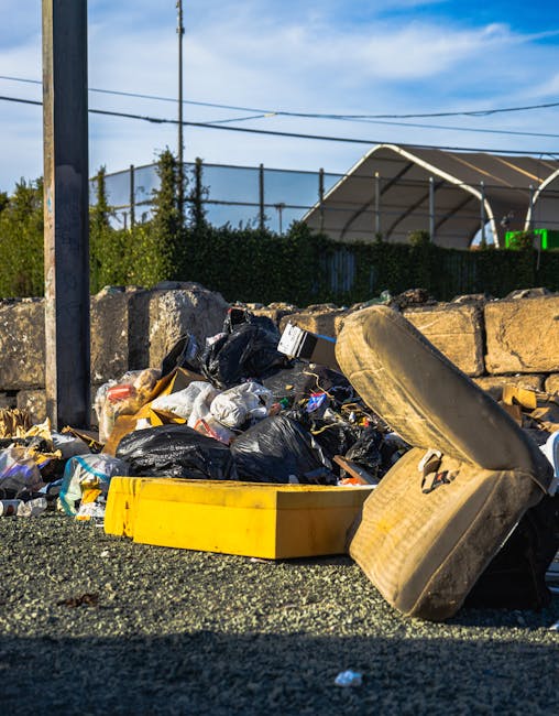A large accumulation of mixed household waste and rubbish bags, including black plastic garbage bags, a yellow plastic tray, and an old, worn car tire placed on a gravel surface, with a stone wall and