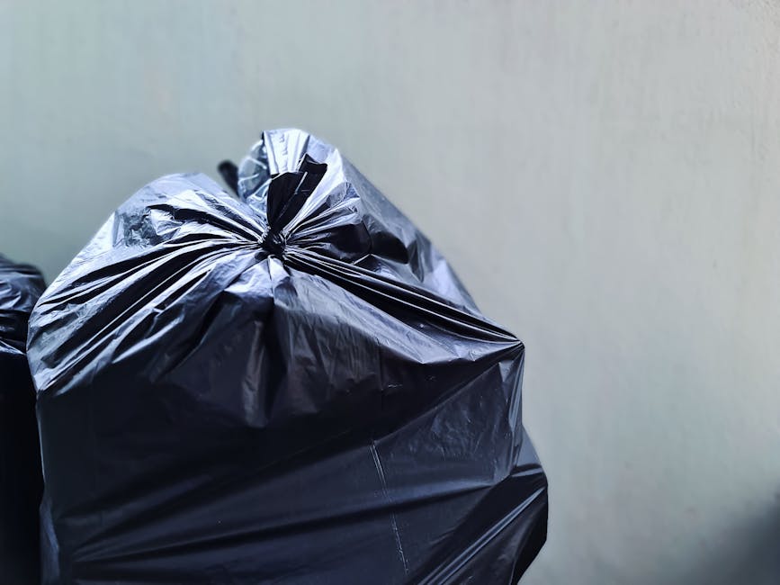 A large accumulation of mixed household waste and rubbish bags, including black plastic garbage bags, a yellow plastic tray, and an old, worn car tire placed on a gravel surface, with a stone wall and