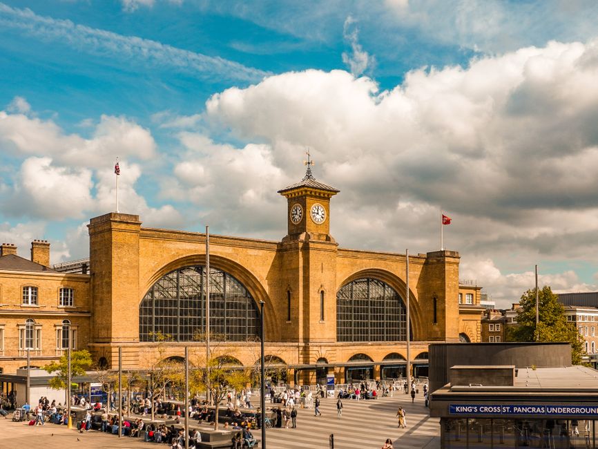 A large historical brick building with arched windows and a central clock tower featuring an ornate spire, situated at Kings Cross station. The foreground shows a busy outdoor area with numerous peopl
