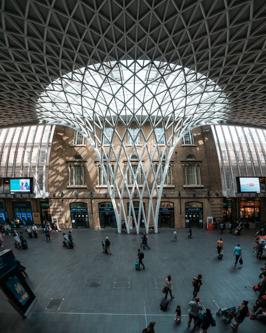 A large historical brick building with arched windows and a central clock tower featuring an ornate spire, situated at Kings Cross station. The foreground shows a busy outdoor area with numerous peopl